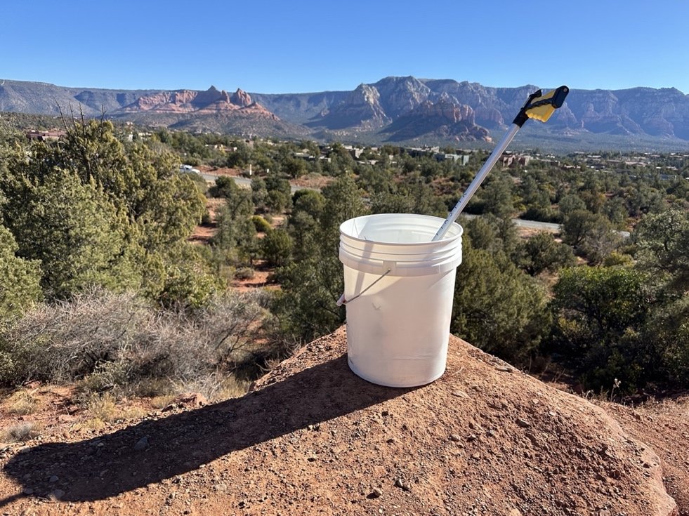 A litter picker and white bucket on a red rock overlook above Sedona, with Cathedral Rock in the distance